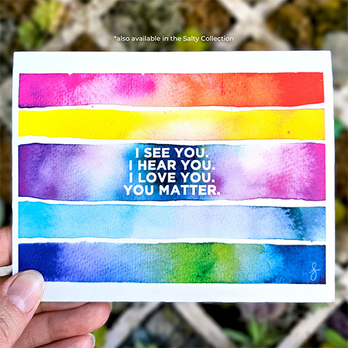 The Pride "You Matter" card is being held against a background of succulents. The card is a watercolor rainbow of colors with the words "I see you. I hear you. I love you. You matter." in white text against the watercolor background.